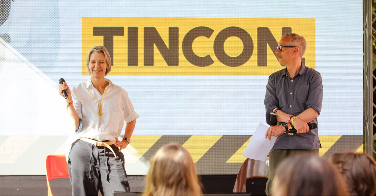 Eine Frau und ein Mann, beide mittleren Alters stehen vor einer Leinwand, auf der TINCON steht.